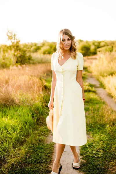 一个年轻漂亮的姑娘 一头金发卷曲 穿着浅黄色的夏装 手中拿着一顶草帽 夕阳西下 在乡间的田野里 人与自然 — 图库照片