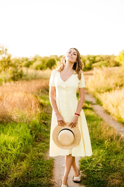 一个年轻漂亮的姑娘 一头金发卷曲 穿着浅黄色的夏装 手中拿着一顶草帽 夕阳西下 在乡间的田野里 人与自然 — 图库照片