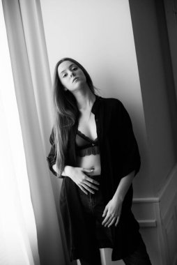 Uzun saçlı, kotlu ve siyah gömlekli çekici genç kız geniş bir pencerede duruyor. 