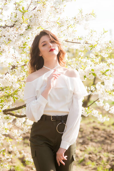Lovely young girl in a white romantic silk blouse in spring in a blooming cherry garden in sunny weather. Spring and Fashion