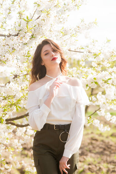Lovely young girl in a white romantic silk blouse in spring in a blooming cherry garden in sunny weather. Spring and Fashion