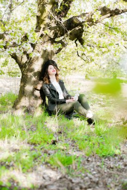 Siyah şapkalı, şık siyah deri ceketli, güneşli bir havada, çiçek açan kiraz bahçesinde elinde çiçek açan bir dalla güzel bir genç kız. Bahar ve Moda