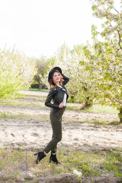 美丽的小女孩 戴着黑色的帽子 穿着时髦的黑色皮夹克 在春天里的一个盛开的樱桃园里 手里拿着一支盛开的樱桃枝 春天与时尚 — 图库照片