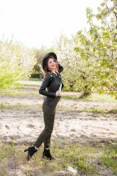 美丽的小女孩 戴着黑色的帽子 穿着时髦的黑色皮夹克 在春天里的一个盛开的樱桃园里 手里拿着一支盛开的樱桃枝 春天与时尚 — 图库照片