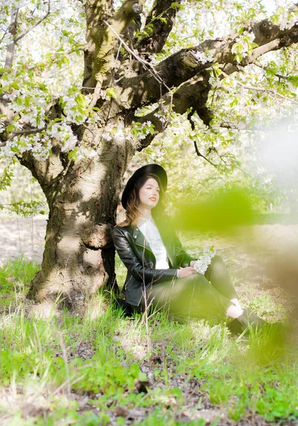 黒い帽子の素敵な若い女の子と彼女の手の中に開花桜の枝と晴れた日の桜の庭で春にファッショナブルな黒い革のジャケット 春とファッション — ストック写真