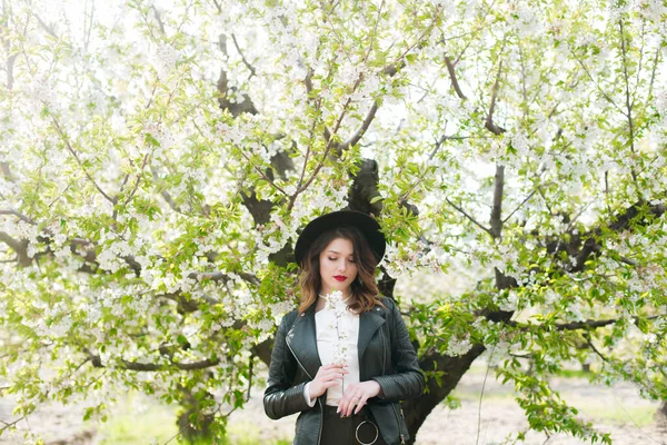 黒い帽子の素敵な若い女の子と彼女の手の中に開花桜の枝と晴れた日の桜の庭で春にファッショナブルな黒い革のジャケット 春とファッション — ストック写真
