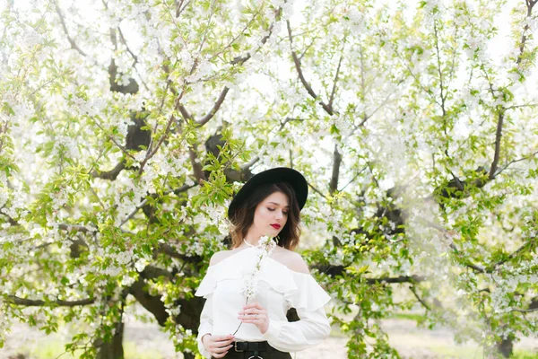 黒い帽子の素敵な若い女の子と彼女の手の中に開花桜の枝と晴れた日の桜の庭で春に白いロマンチックなシルクのブラウス 春とファッション — ストック写真