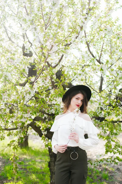 黒い帽子の素敵な若い女の子と彼女の手の中に開花桜の枝と晴れた日の桜の庭で春に白いロマンチックなシルクのブラウス 春とファッション — ストック写真