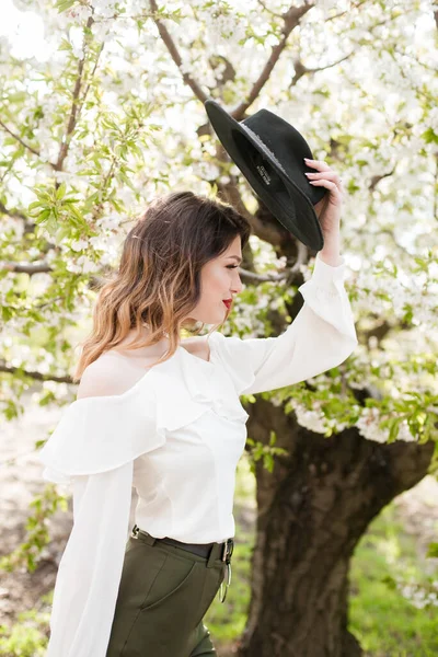 美丽的小女孩 戴着黑色的帽子 穿着白色的浪漫丝绸衬衫 在春天里的一个盛开的樱桃园里 手里拿着一支盛开的樱花枝头 春天与时尚 — 图库照片