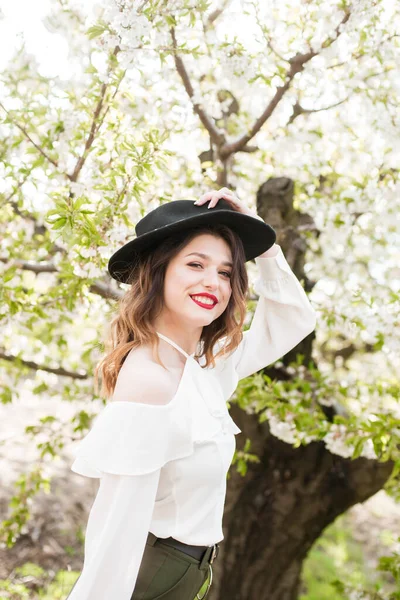 美丽的小女孩 戴着黑色的帽子 穿着白色的浪漫丝绸衬衫 在春天里的一个盛开的樱桃园里 手里拿着一支盛开的樱花枝头 春天与时尚 — 图库照片