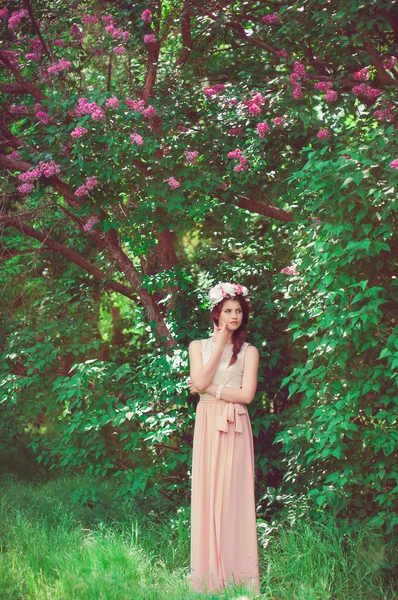 Mooi meisje in een lange jurk en een krans van bloemen van lila bush — Stockfoto