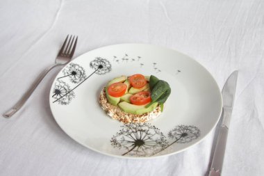 Beyaz bir arka planda, beyaz bir tabakta üzerinde avokado ve vişneli domates dilimleri olan yuvarlak bir somun var.