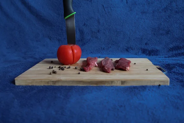 木の板の上には 生の肉 スパイス トマトの破片があり ナイフが青い背景に突き刺さっています — ストック写真