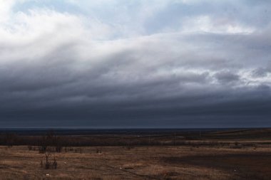 Sert manzara: Kuru çimenler ve karla kaplı kahverengi tepelerin üzerinde kara bulutlar asılı.