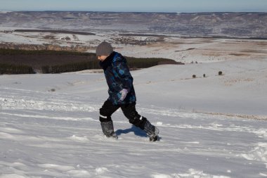 Bir çocuk karla kaplı bir tarlada koşuyor, arka planda dağlar ve mavi gökyüzü görünüyor.