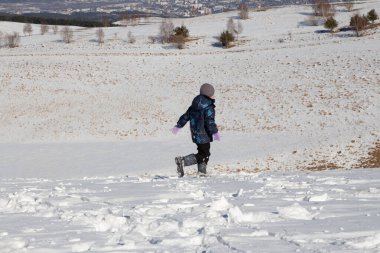 Bir çocuk karla kaplı bir tarlada koşuyor, arka planda dağlar ve mavi gökyüzü görünüyor.