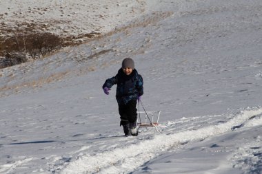 Kışın giyinmiş bir çocuk karlı bir tarlada koşar ve arkasından bir kızak çeker. 