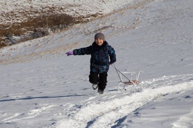 Kışın giyinmiş bir çocuk karlı bir tarlada koşar ve arkasından bir kızak çeker. 