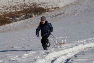 Kışın giyinmiş bir çocuk karlı bir tarlada koşar ve arkasından bir kızak çeker. 