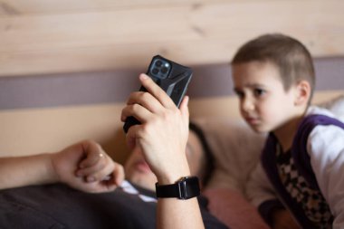Bir adam kanepede uzanıyor ve elinde bir telefon tutuyor. Yanında bir çocuk telefona bakıyor.