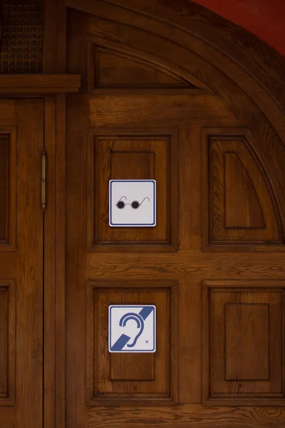 A sign hangs on a wooden door: black glasses on a white background - blind pedestrians, and a blue stylized ear - deaf