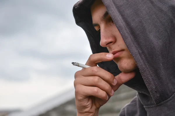 若有所思和担心十几岁的男孩，与黑色连帽衫是吸烟 cig — 图库照片