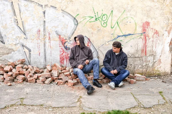 Dos vándalos callejeros o raperos de pie contra una pared pintada de graffiti se preparan para fumar un cigarrillo — Foto de Stock