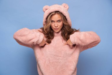 Huysuz, öfkeli bir kızın portresi kameranın merceğine bakıyor mavi arka planda ayı kıyafeti giymiş parmaklarıyla kendine dokunuyor.