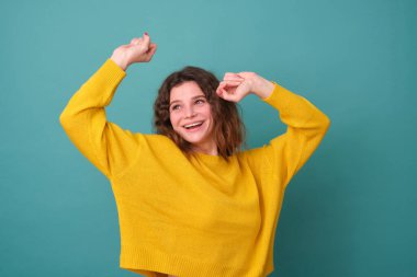 Kıvırcık saçlı neşeli bir manken, neşeli, sarı bir kazak giyiyor, geniş bir yelpazede gülümsüyor, eğleniyor ve mavi arka planda izole edilmiş müziğin ritmine göre hareket ediyor.