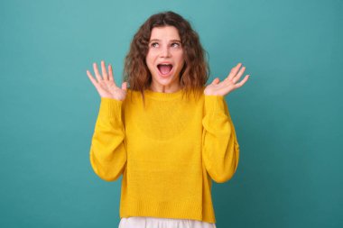 Kıvırcık saçlı tatlı bir kız eğleniyor, ellerini kaldırıyor, geniş bir gülümseme sergiliyor, komik bir şey görüyor, mavi arka planda izole edilmiş sarı bir kazak giyiyor.