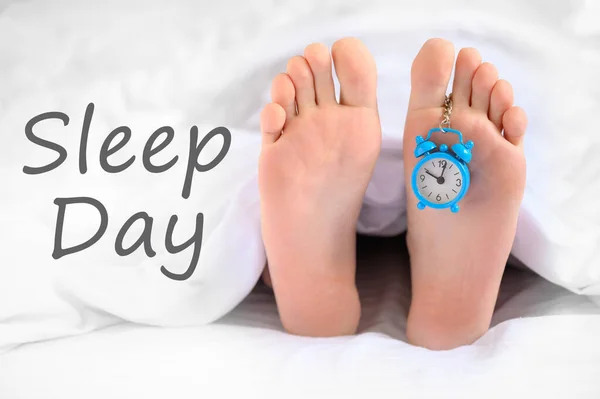 Sleep day. A man lies in bed, an alarm clock hangs on his leg, the concept of laziness, world sleep day, time to get up.