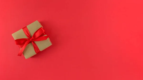 Caja de regalo envuelta en un papel artesanal con cinta roja y lazo sobre fondo rojo. Monocromo plano festivo con espacio de copia. — Foto de Stock