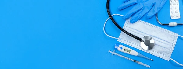 Stethoscope, face mask, medical syringe, thermometer, pills blister and medical gloves on blue background. Flat lay banner with copy space. — Stock Photo, Image