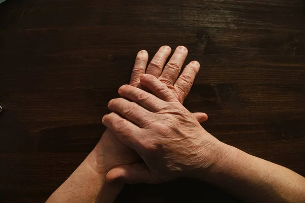 Hands and arms of old man lying on top of wooden table surface. Directly above.