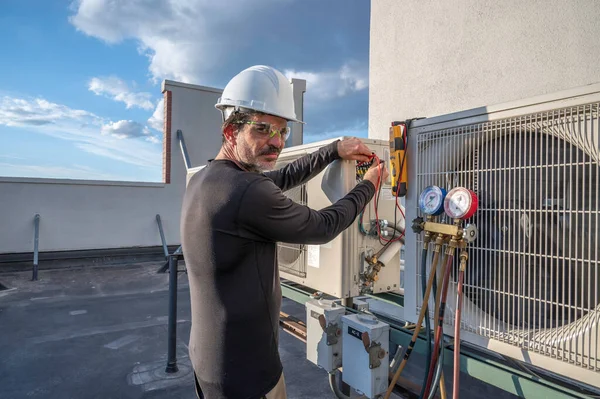 HVAC teknisyeni kameraya bakıyor, voltajı küçük bir havalandırma sistemi üzerinde test ediyor.
