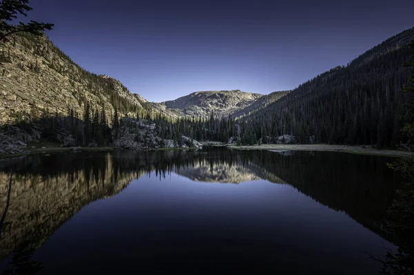 Dağlar ve ağaçlar, nadiren ziyaret edilen bir vadide bulunan bu uzak dağlık alp gölünün sakin sularını yansıtıyor: Rocky Dağı Ulusal Parkı, Colorado, ABD