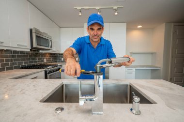 Mavi üniformalı tesisatçı mutfak musluğunu tamir etmek ya da kurmak için pense kullanıyor.