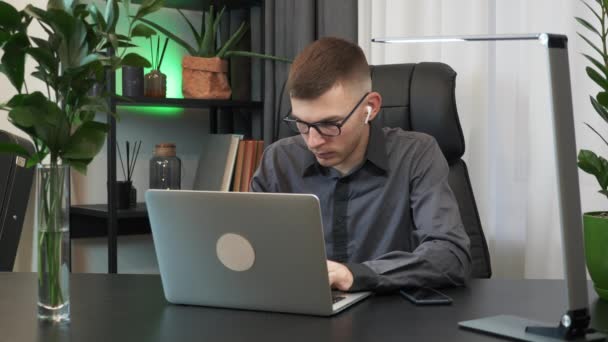 젊은 사업가가 노트북 컴퓨터로 키보드를 치고 화면을 보고 인터넷 뉴스를 읽고 있습니다. 안경과 이어폰을 착용하고 있는 남자가 직장에서 현대 사무실에서 일하고 있다 — 비디오