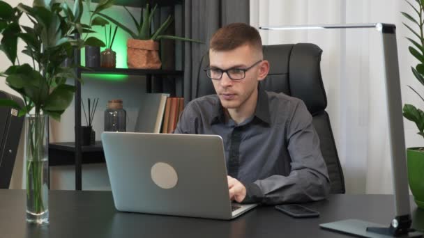 Мотивированный молодой человек закрывает ноутбук и заканчивает работу за рабочим столом вечером. Бизнесмен в очках заканчивает работу над блокнотом в офисе. Концепция бизнеса — стоковое видео