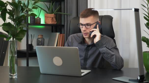 Молодой бизнесмен сидит за рабочим столом в своем современном офисе и разговаривает по смартфону с партнерами. Многозадачный человек работает над ноутбуком и разговаривает по телефону. Мужчина многозадачен на работе — стоковое видео