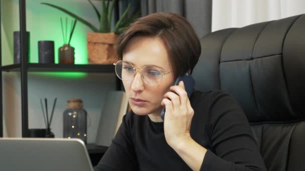 Retrato de la joven mujer de negocios hablando en el teléfono inteligente frente a la computadora portátil en la oficina moderna. Mujer en gafas está trabajando en el ordenador, mirando la pantalla y hablando con los socios en el teléfono — Vídeos de Stock
