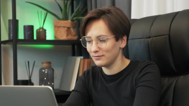 Mujer sonriente enfocada en gafas está trabajando en el ordenador portátil en la oficina. La empresaria está usando un cuaderno en el escritorio del lugar de trabajo. Atractiva mujer trabaja en el ordenador portátil en la oficina en casa por la noche — Vídeos de Stock