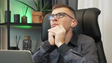 Düşünceli, ciddi bir genç adam elinde dizüstü bilgisayarla modern ofis masasında oturuyor ve düşüncelere dalmış, sorunlu çözüm fikirleri düşünüyor. Adam mali sorunlardan endişeleniyor.