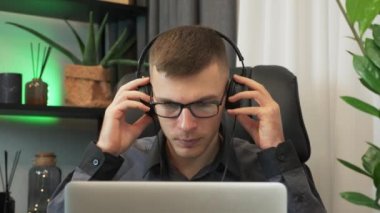 Genç erkek serbest çalışan kulaklık takar ve ofisteki dizüstü bilgisayarda çalışmaya başlar. Gülümseyen adam kulaklıktan müzik dinliyor ve modern çalışma masasında defter üzerinde çalışıyor.