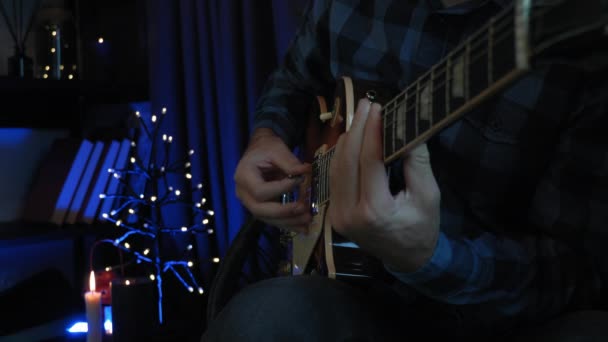 O guitarrista profissional toca balada lírica romântica na guitarra elétrica na sala de estar escura com luzes no fundo. Homem está tocando na guitarra sentado no sofá no apartamento moderno — Vídeo de Stock