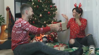 Kadın Noel hediyesi alıyor, eğleniyor ve sallanıyor. Mutlu çift evde bayram kutluyor. Dişi, hediyesini açacağı için heyecanlı. Hediye kutusunu sallıyor ve gülümsüyor.