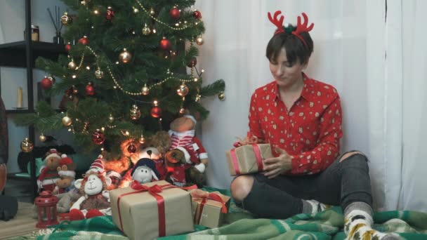 Encantadora mujer sonriente está envolviendo y decorando regalos de Navidad mientras se sienta cerca del árbol de Navidad en casa por la noche, preparándose para el Año Nuevo 2021 y las vacaciones de Navidad — Vídeo de stock