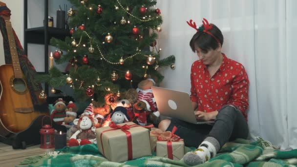 Mujer morena atractiva en ropa de fiesta de Navidad está sentado cerca del árbol de Navidad y trabajando en la computadora en la noche de Año Nuevo. Hay muchas cajas de regalos debajo de los árboles. Concepto espíritu navideño — Vídeo de stock