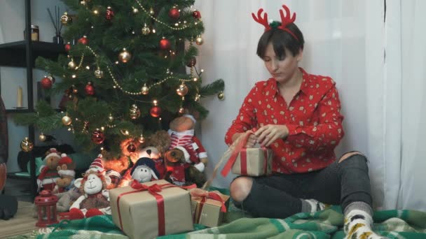Espíritu navideño, vacaciones y concepto de celebraciones. Joven alegre en ropa de fiesta está envolviendo regalos de Navidad en papel y lazo rojo. Mujer decorando la caja de regalo de Navidad. Feliz Año Nuevo — Vídeo de stock