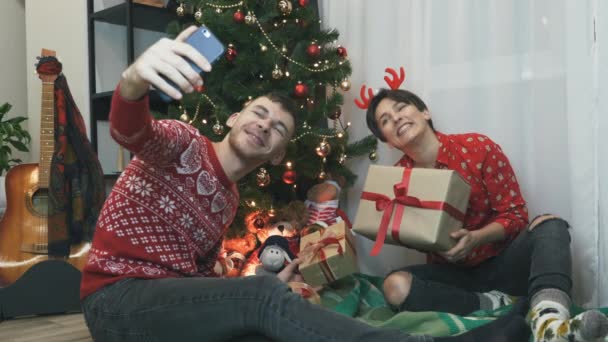 Ung familj njuter och firar julhelgen tillsammans hemma. Kvinna och man sitter nära granen och gör selfies. Julstämning, firande koncept — Stockvideo